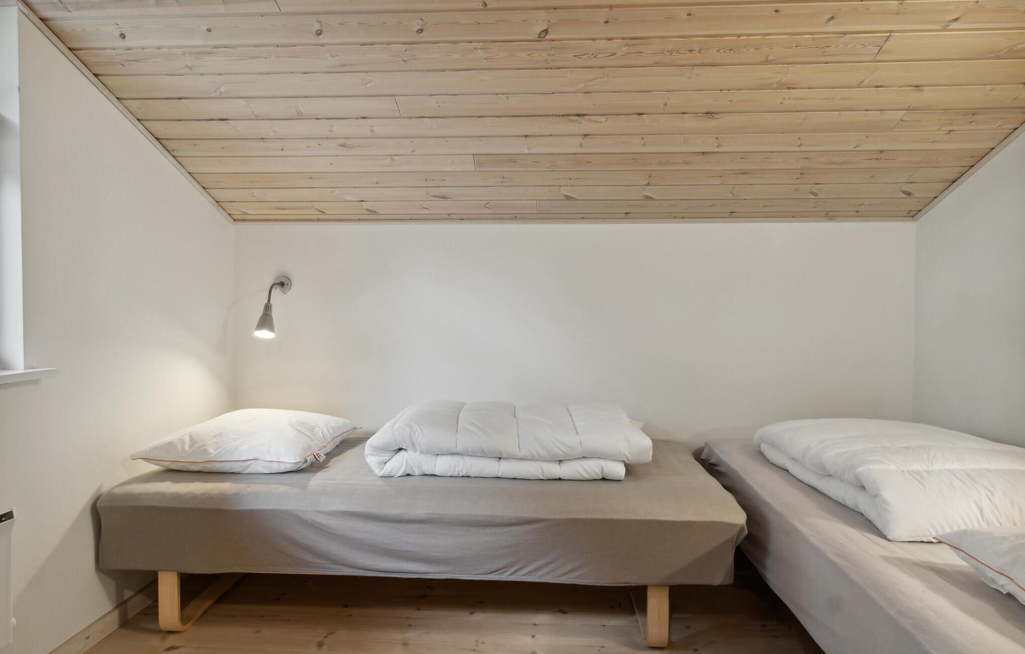 A small, single-room with a sloped ceiling, two beds, a wall-mounted light, and a window.