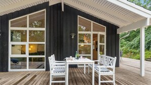 Un espacio de estar al aire libre cubierto con una mesa y dos sillas blancas, ventanas amplias y una terraza de madera.
