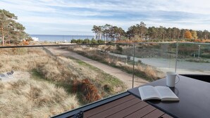 Vista desde un balcón con un libro y una taza en una mesa, con vista a un sendero costero con árboles y una masa de agua.