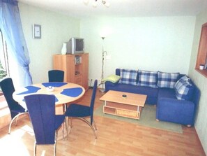 Ein Wohnzimmer mit blauem Sofa, einem Holztisch, einem Esstisch mit Stühlen, einem Fernseher auf einer Kommode und einer Lampe.