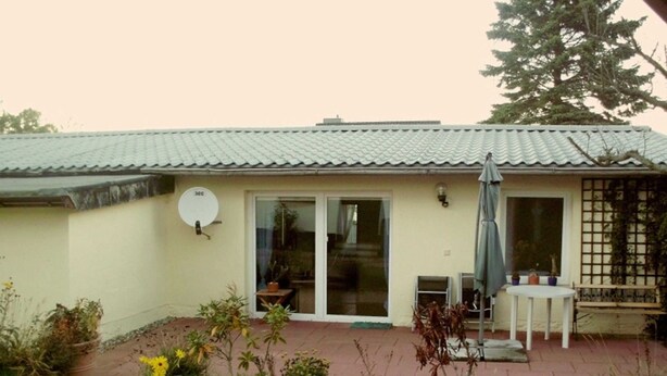 Una casa con tetto in tegole, antenna parabolica, porta scorrevole in vetro, patio con tavolo e sedie, e un piccolo giardino con piante e fiori.