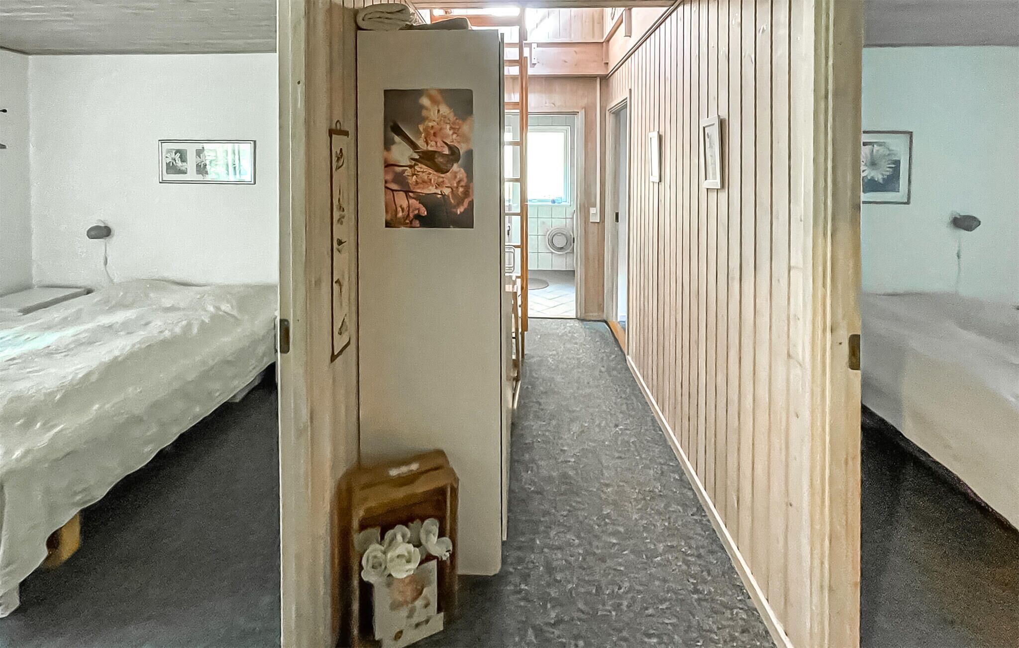 A corridor with wooden paneling, a bed on the left, a door with a picture, and a small table with flowers.