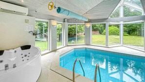 En moderne swimmingpool indendørs med krystalklart vand, et boblebad og store vinduer med udsigt til en velanlagt have.
