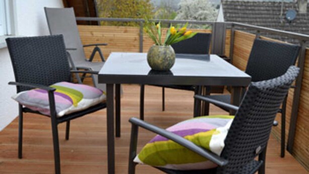 A balcony with a table and chairs, a vase with flowers, and a view of trees and buildings. - Himmel-Blick - Vacation Apartment Himmel-Blick (Pünderich)
