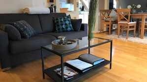 A modern living room with a dark grey sofa, a glass coffee table, and a dining area with wooden chairs and a white tablecloth. - Himmel-Blick - Vacation Apartment Himmel-Blick (Pünderich)