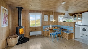 A wooden cabin interior with a central fireplace, a dining area with blue chairs, and a kitchen with a washing machine. - Cozy home in Frøstrup with kitchen (Frøstrup)