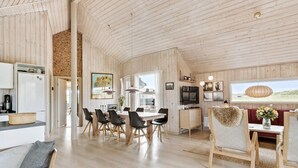 A spacious room with a wooden ceiling, a dining area with a table and chairs, a kitchen area with a refrigerator, and a large window offering a view of the outdoors. - Cozy home in Hvide Sande with sauna (Hvide Sande)