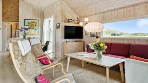 A cozy living room with wooden walls, a flat-screen TV, a red sofa, and a view of the countryside. - Cozy home in Hvide Sande with sauna (Hvide Sande)