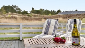 A wooden deck with a table, two chairs, a bottle of wine, and a bunch of grapes.