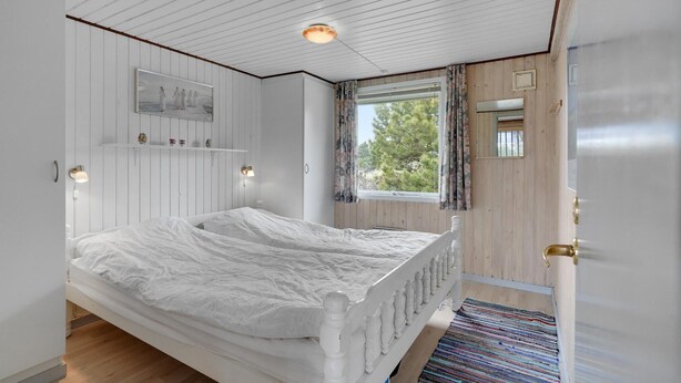 A bedroom with a bed, a window with curtains, a wooden ceiling, and a small shelf with decorative items.