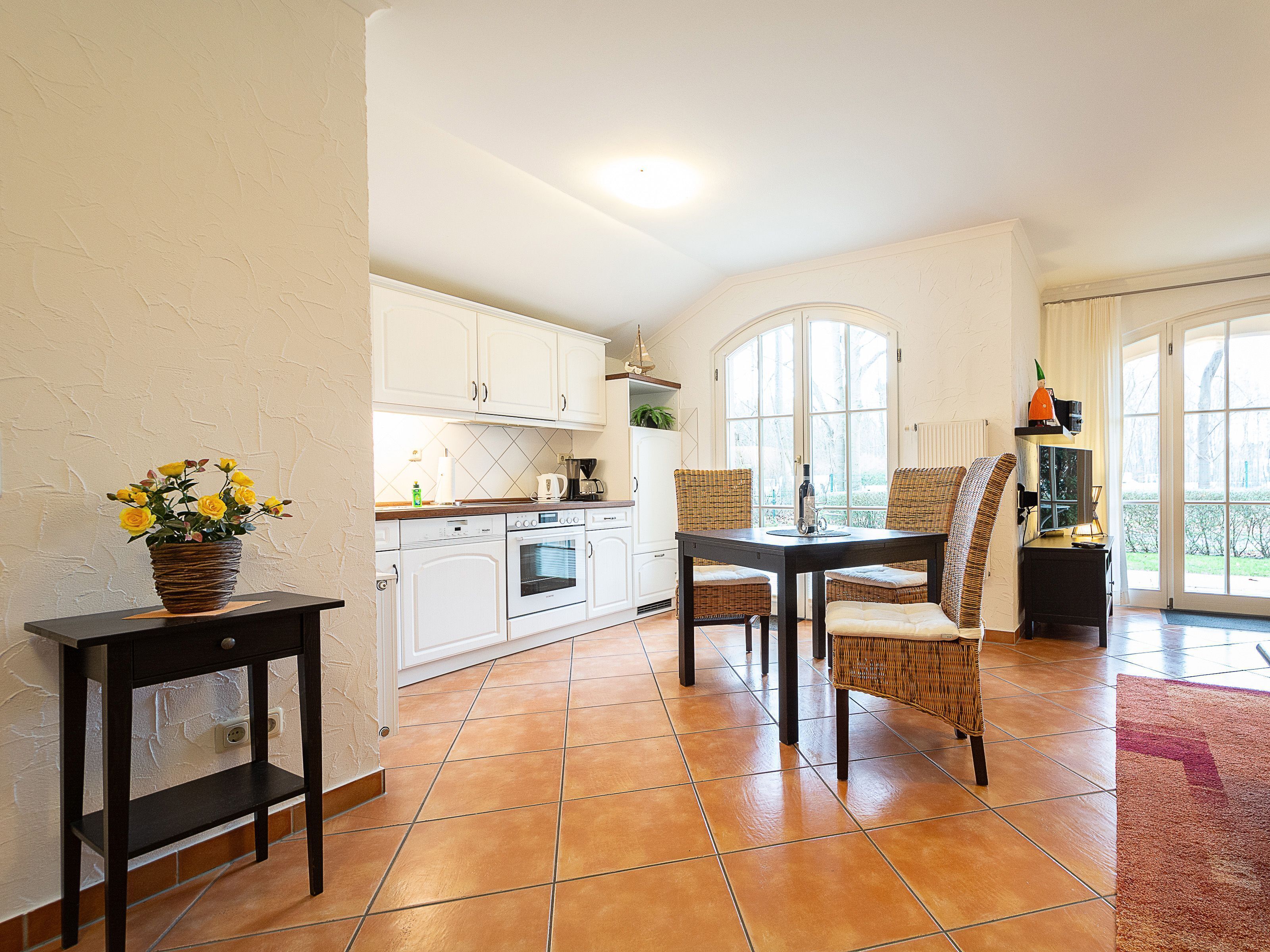 Une cuisine avec des meubles blancs, une table à manger avec des chaises et une plante en pot posée sur une table d’appoint.