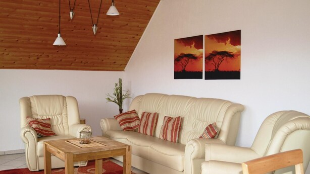 Un salon avec un plafond en bois, un canapé beige, deux fauteuils, une table basse en bois et un tapis rouge à motifs.