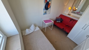 Um quarto pequeno com uma cama, uma mesa, um sofá vermelho com caracteres chineses, um quadro na parede e um guarda-roupa.