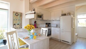 Cocina moderna con muebles blancos, microondas integrado y pileta de acero inoxidable. Hay un pequeño comedor con una silla blanca y una mesa, y un jarrón con girasoles en la encimera.