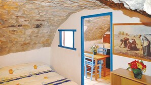 A bedroom with a stone wall, a bed with floral bedding, a small table with a chair, a painting, and a vase with flowers. - 1 bedroom nice apartment in La Beaume (La Beaume)