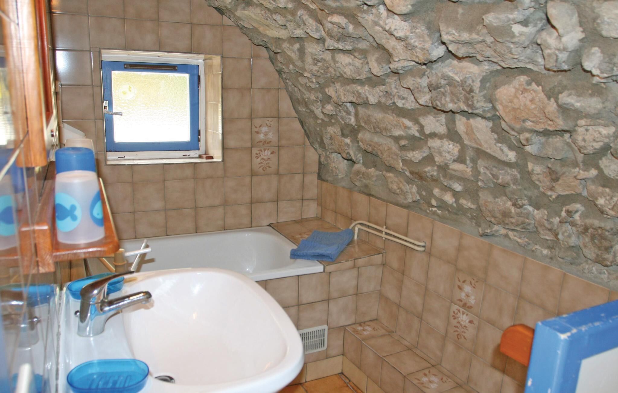 Ein Badezimmer mit einem weißen Waschbecken, einer Badewanne und einer Duschkabine. Über dem Waschbecken befindet sich ein Fenster und an einer Seite eine Steinwand.