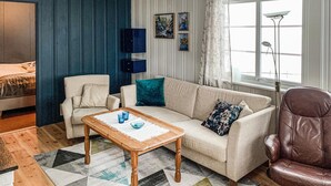 A living room with a sofa, armchair, and coffee table. There is a window with a view, a lamp, and wall-mounted artwork.