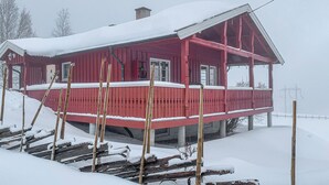 A red wooden house with a snow-covered roof and a balcony, surrounded by snow.