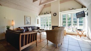 A cozy living room with a wooden ceiling, a sofa, a wicker chair, a fireplace, and large windows offering a view of greenery.
