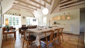 A spacious dining area with a long table and wooden chairs, a cozy seating area with a sofa and chairs, and a kitchen area with cabinets and a hanging light fixture.