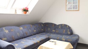 Ein Zimmer mit einem blauen Sofa, einem kleinen Tisch mit einem Glas, einem gerahmten Bild an der Wand und einem Fenster mit einer Pflanze.