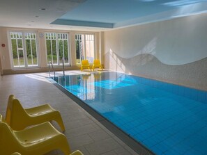 An indoor swimming pool with clear blue water, yellow chairs, and large windows allowing natural light.