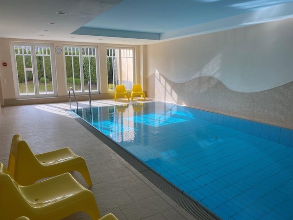 An indoor swimming pool with clear blue water, yellow chairs, and large windows allowing natural light. - (26) 2-room apartment Ostseeallee - apartment house dune castle (Kühlungsborn)