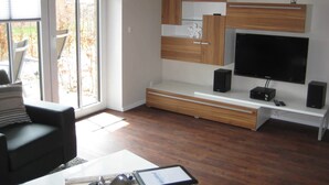 A modern living room with a large TV, wooden cabinets, and a sliding glass door to the outside. - 2-room 001 Amrum Terr. Dish washer. (BC 5) - BUE - Apartment house Westwind (Büsum)