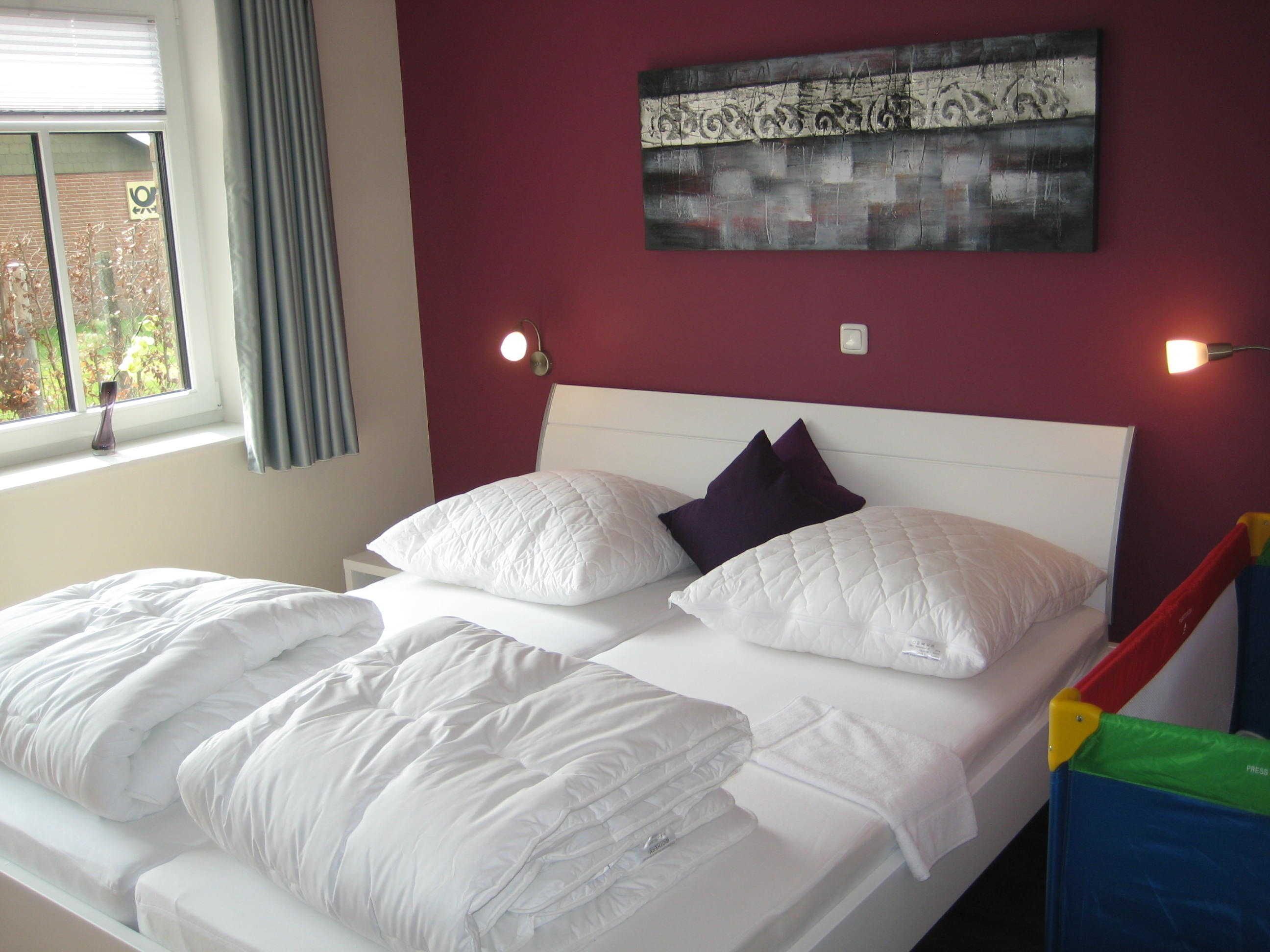 A neatly made bed with white bedding and two pillows, a window with curtains, and a wall with a painting.
