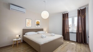 Un dormitorio con una cama, una mesita de noche con lámpara, dos cuadros enmarcados y un ventanal con cortinas.