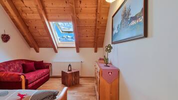 Une pièce chaleureuse avec un plafond en bois incliné, un canapé rouge, une petite table sur laquelle est posée une lanterne, un radiateur et un tableau accroché au mur.