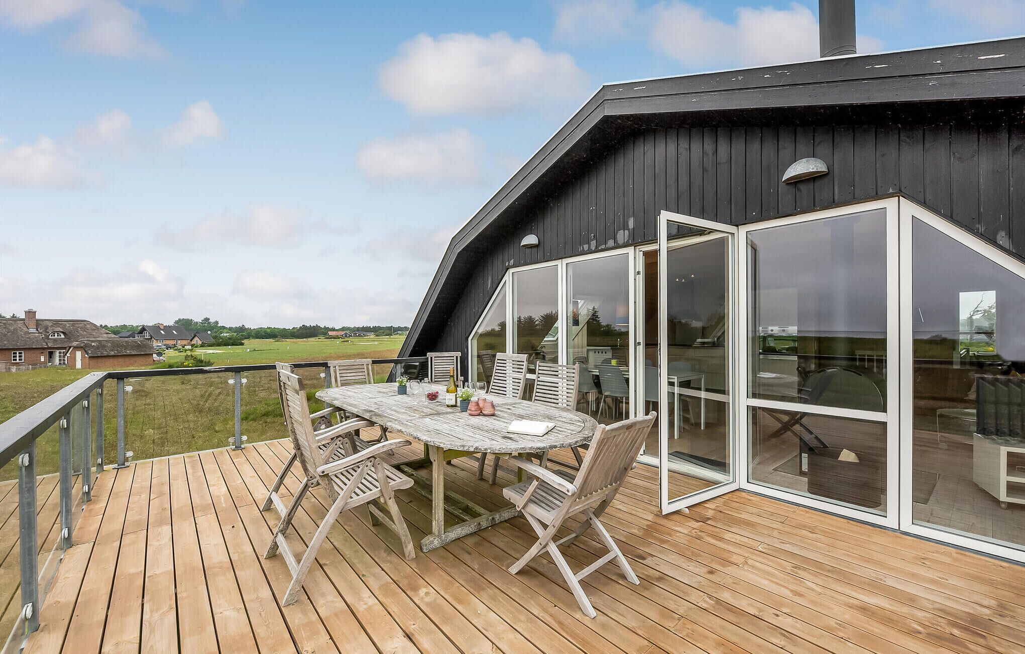 En træterrasse med et bord og stole, en glasdør der fører til et værelse, og udsigt over landskabet.