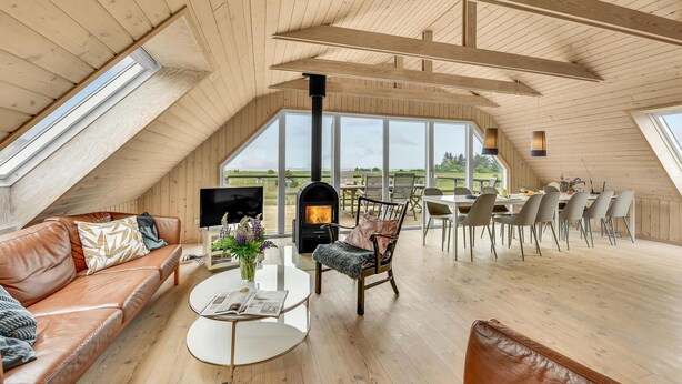 A cozy loft-style room with a wooden ceiling, a fireplace, a sofa, a dining table, and large windows offering a view of the outdoors. - Amazing home in Skjern with sauna (Skjern)