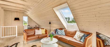 Un acogedor espacio tipo loft con techo inclinado, paredes de madera, un sofá de cuero marrón, una mesita de café blanca y una pequeña mesita auxiliar.