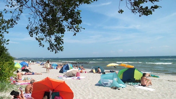 Een strandscène met mensen die ontspannen onder parasols en tenten, een heldere lucht en de oceaan op de achtergrond.