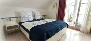 A bedroom with a bed, two pillows, a nightstand, a window with curtains, and a radiator.