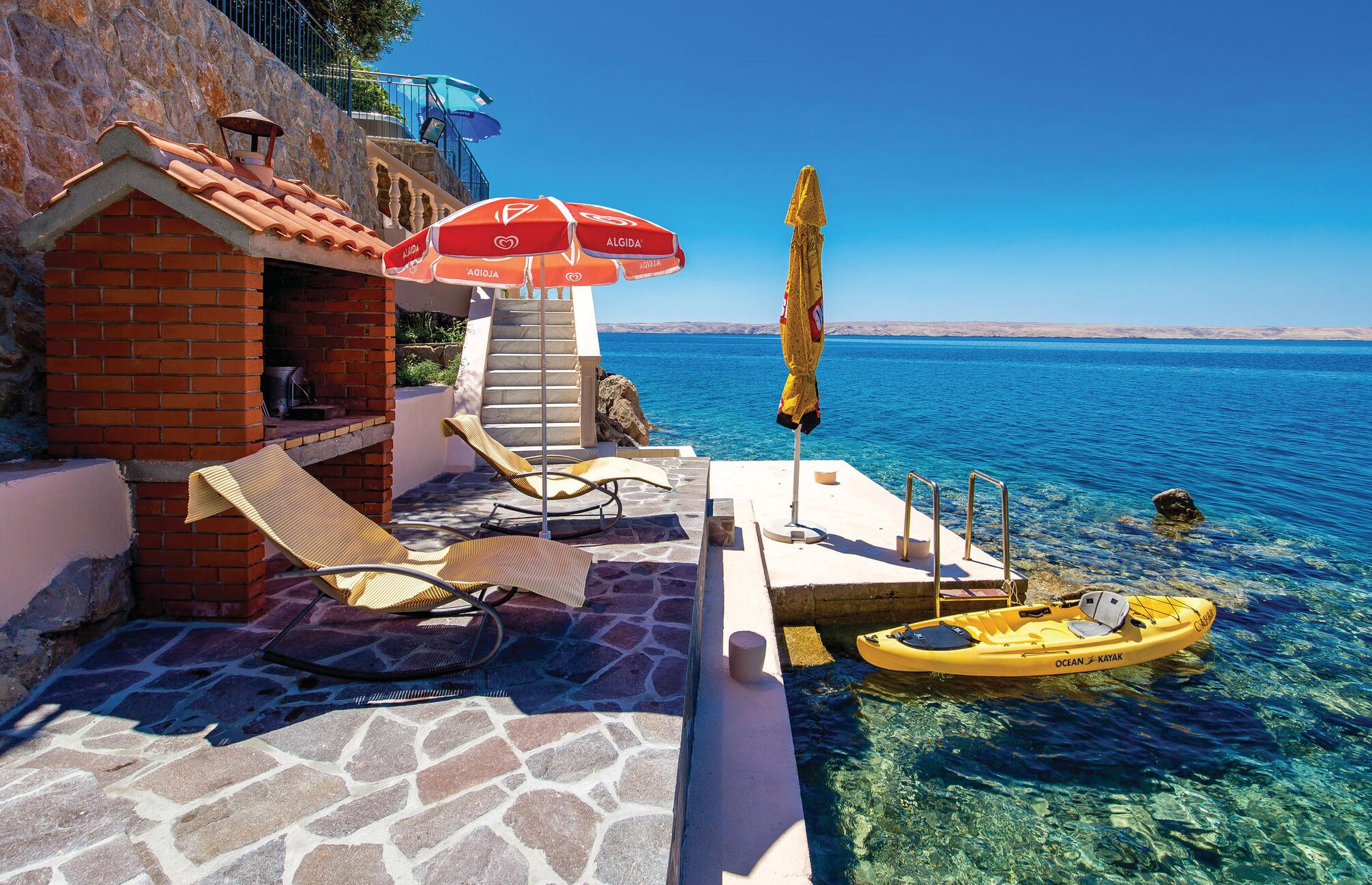 Terraço costeiro com cadeiras de sol, um guarda-sol vermelho e um caiaque junto ao mar.