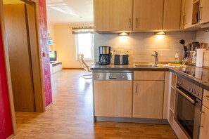 A compact kitchen with wooden cabinets, a dishwasher, a coffee maker, and a stove. - Apartment 1 - Apartments in Blu Hus (Kröslin)