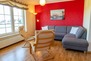 A living room with a red wall, a grey sofa, a wooden chair, and a window with curtains. - Apartment 1 - Apartments in Blu Hus (Kröslin)