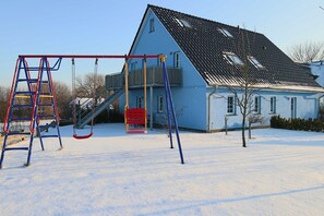 A snow-covered playground with swings and a ladder, a blue house with a balcony, and a clear sky.