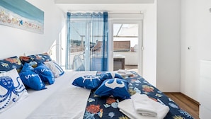 A bedroom with a bed featuring a blue and white nautical-themed bedspread, multiple pillows, and a view of a cityscape through a window with blue curtains. - Lovely home in Murter with WiFi (Murter)