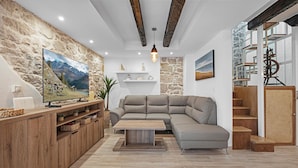A living room with a stone wall, a large flat-screen TV, a grey sectional sofa, a wooden coffee table, and a wooden cabinet with storage. - Lovely home in Murter with WiFi (Murter)