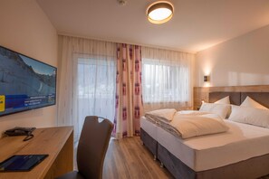 A hotel room with a bed, a desk with a phone and tablet, a chair, and a large window with curtains. - Double room Hochzillertal 2 - Aparthotel **** AlpTirol (Kaltenbach)