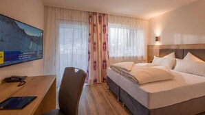 A hotel room with a bed, a desk with a phone and tablet, a chair, and a large window with curtains. - Double room Hochzillertal 2 - Aparthotel **** AlpTirol (Kaltenbach)