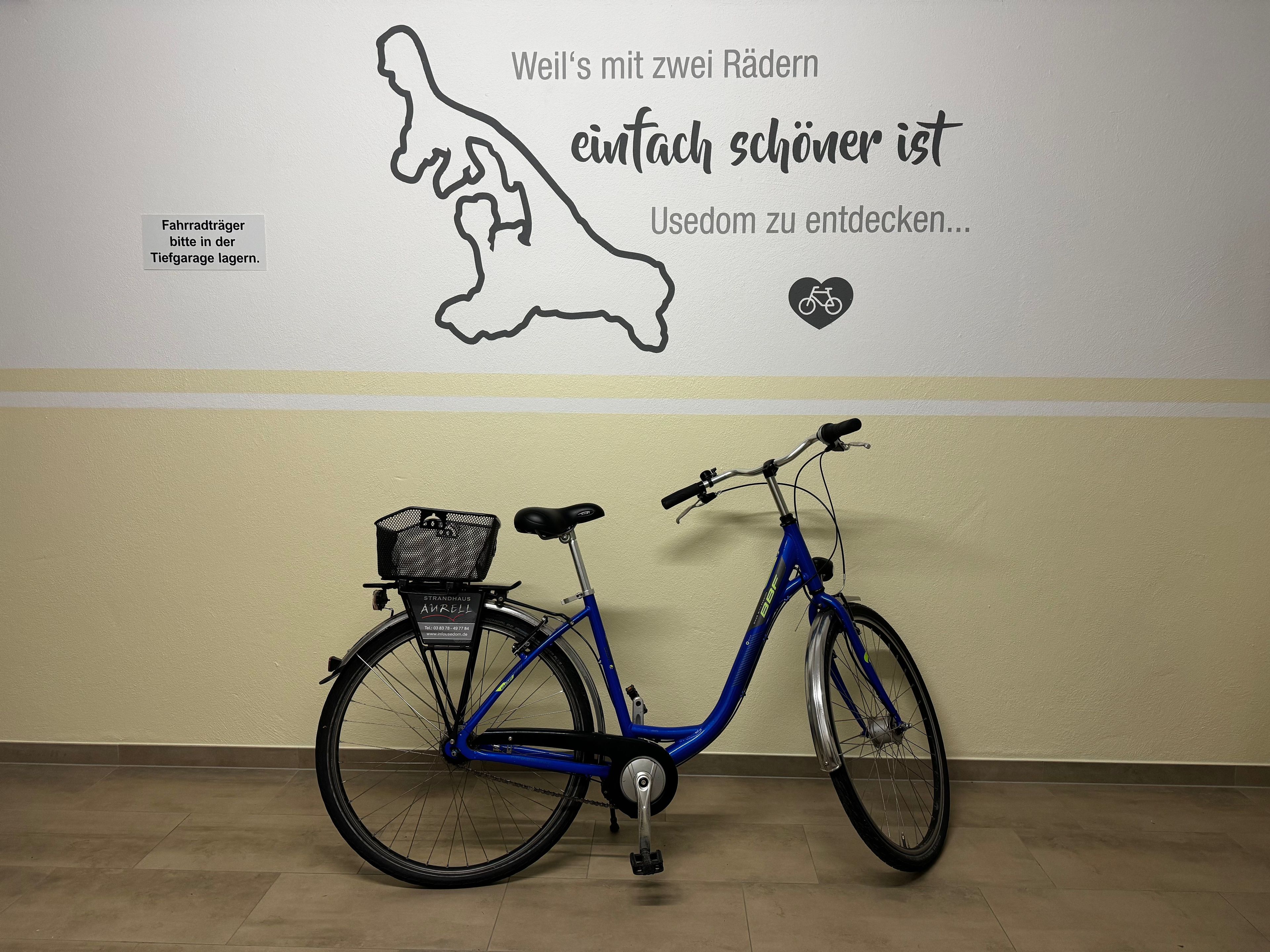 Un vélo bleu avec un panier et un mur orné d’une citation allemande et d’un panneau indiquant : « Veuillez garer les porte-vélos dans le parking souterrain. »