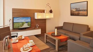 Une chambre d’hôtel moderne avec une tête de lit en bois, un téléviseur à écran plat, un canapé, une table à manger agrémentée d’un chemin de table rouge et un tableau au mur.