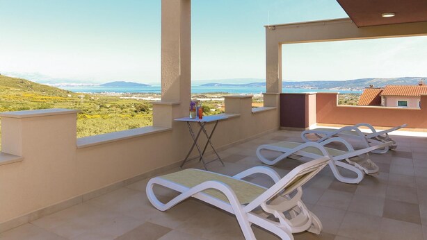 Ein Balkon mit weißen Liegestühlen, einem kleinen Tisch und Blick auf das Meer und die Hügel.