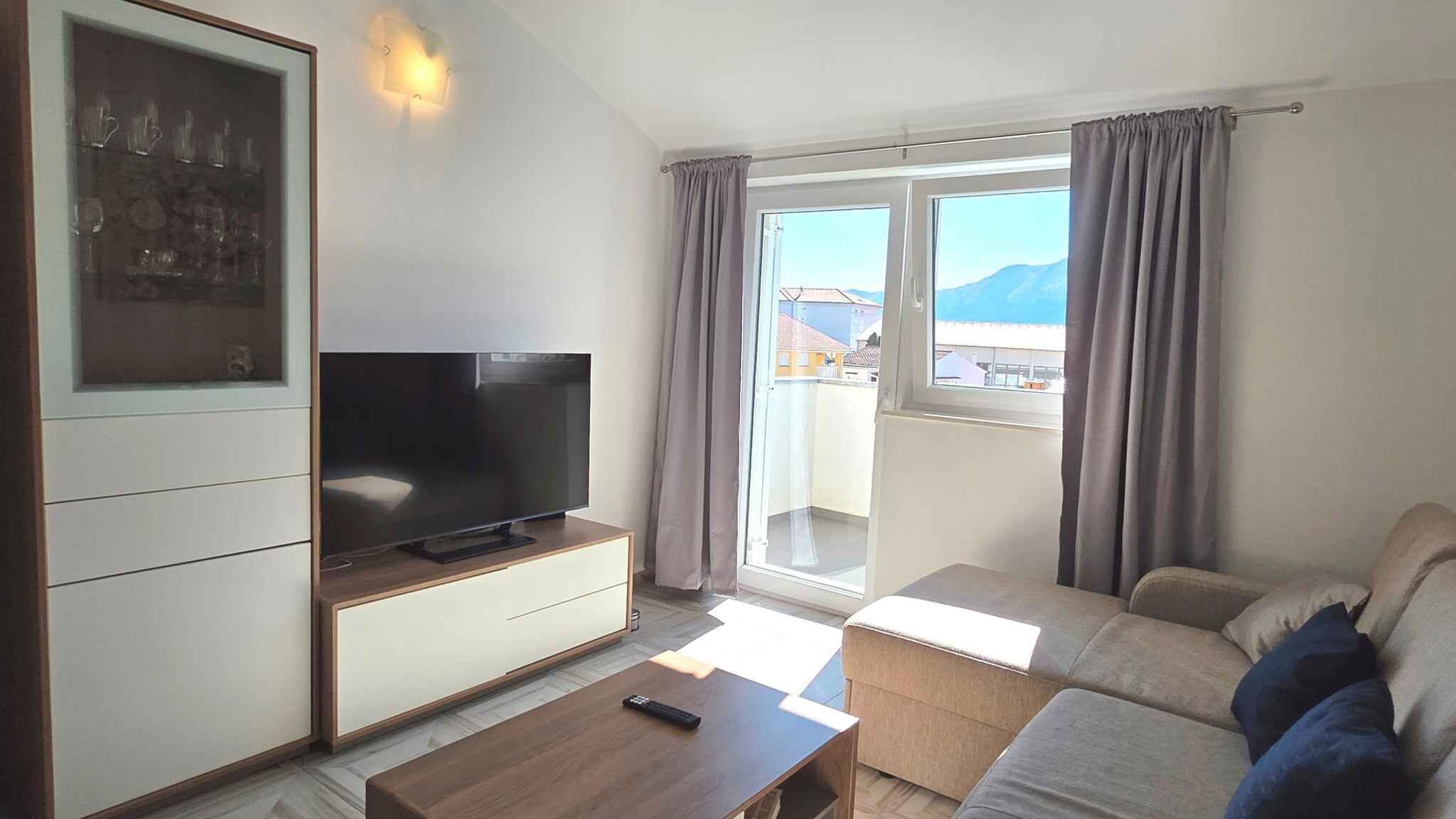 A living room with a flat-screen TV, a sofa, a coffee table, and a balcony with curtains.