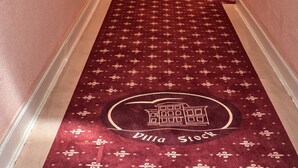 A hallway with a red carpet featuring a building logo, white double doors, and wall-mounted lights.