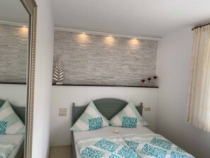 Ein Schlafzimmer mit einem Bett, einem Spiegel, einer Wandablage mit Dekoration und einem Fenster mit Vorhängen.